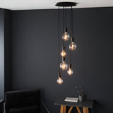 Marlowe 6 Light Pendant in matt black metal, six exposed bulbs on staggered black fabric flex drops, shown against a dark wall above a side table.