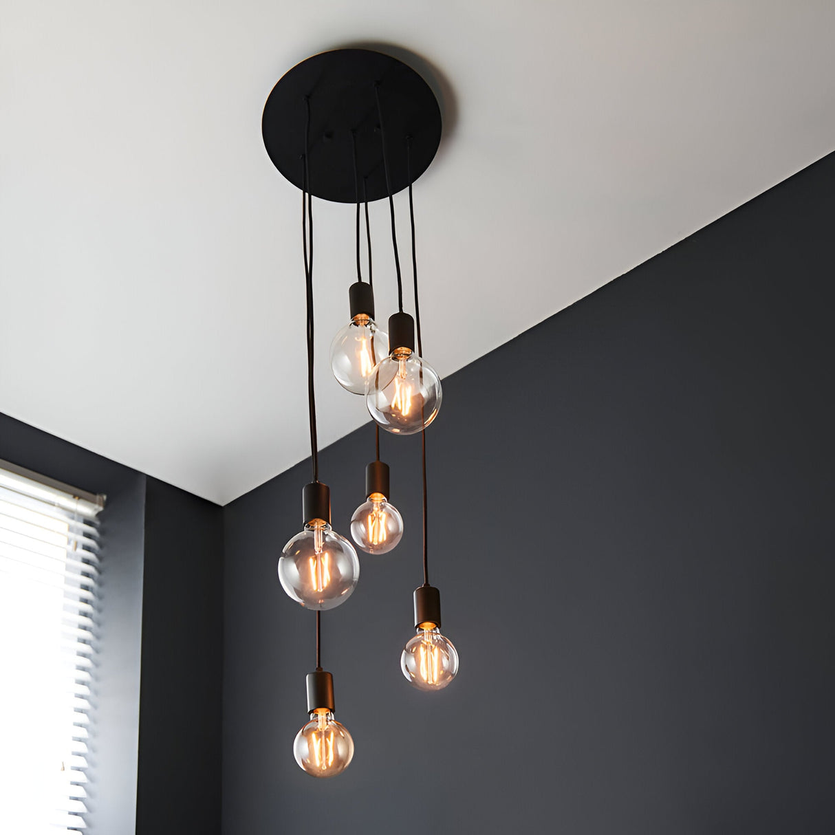 Marlowe 6 Light Pendant in matt black metal, staggered fabric flex drops with exposed globe bulbs, shown against a dark sloped ceiling and grey wall.