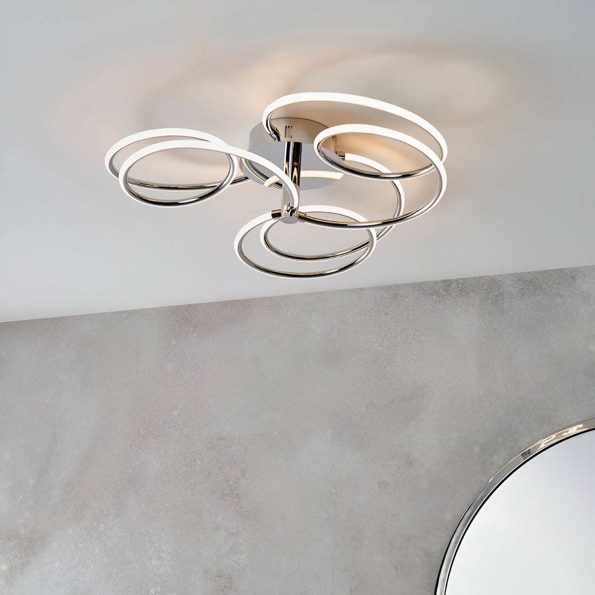 Endon Ravel LED semi-flush ceiling light in chrome, with looping ring-shaped arms radiating from a round base, shown on a white ceiling above a stone wall.