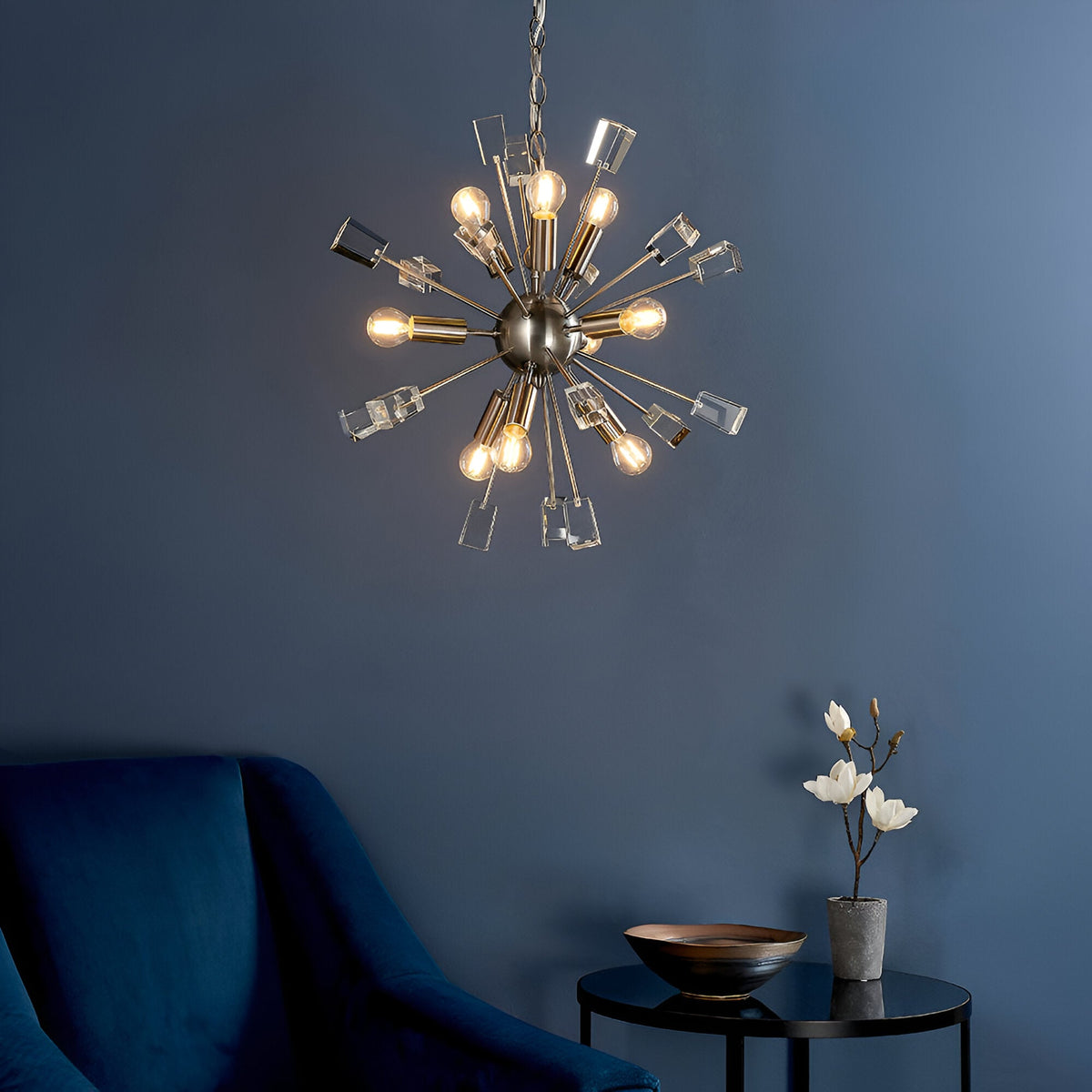 Carlgrove 9 Light Pendant in satin nickel, sputnik-style rods with clear crystal accents, shown lit above a navy armchair in a living room setting.