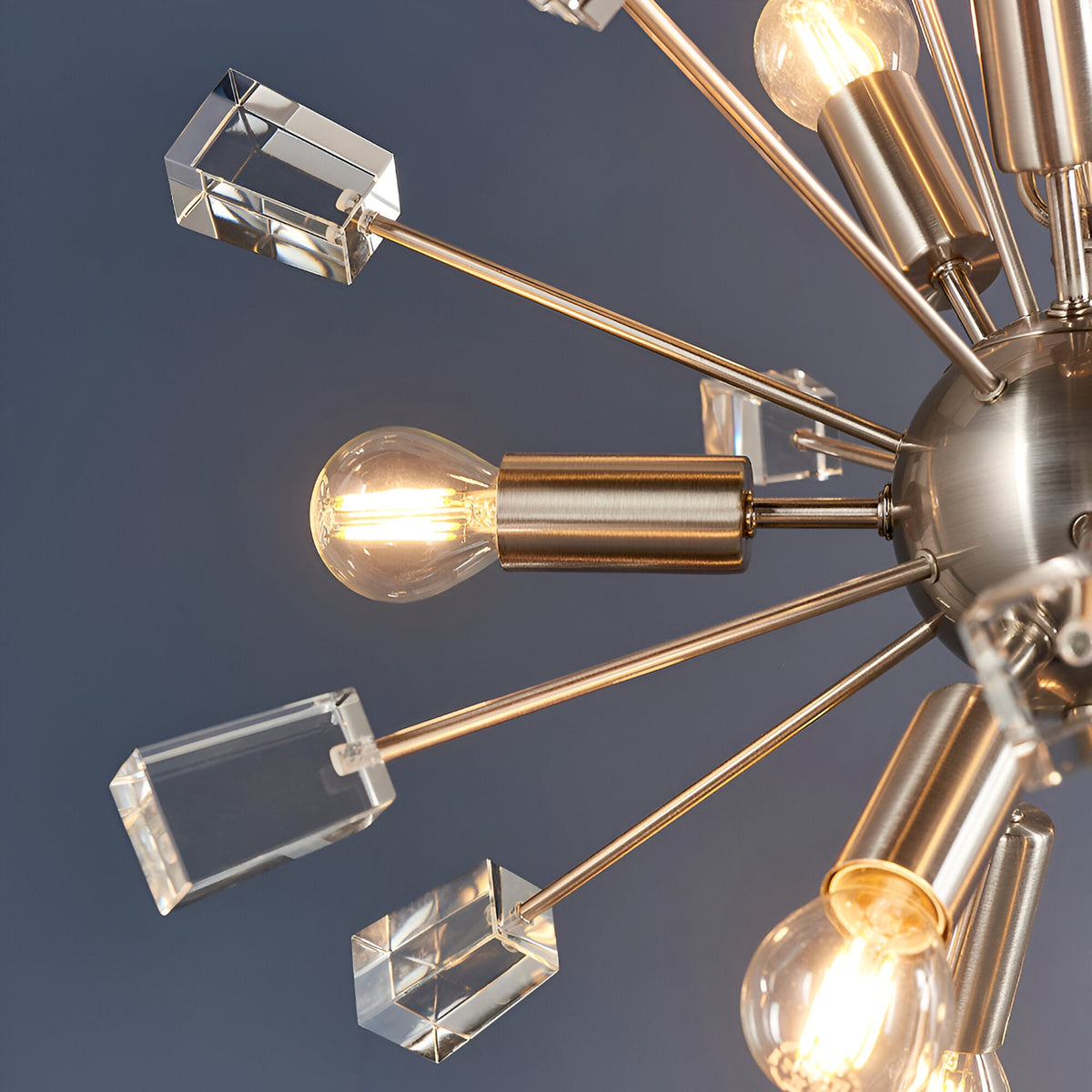 Close-up of the Carlgrove 9-Light Pendant in satin nickel with radiating metal rods, clear crystal glass blocks, and exposed bulbs against a grey background.
