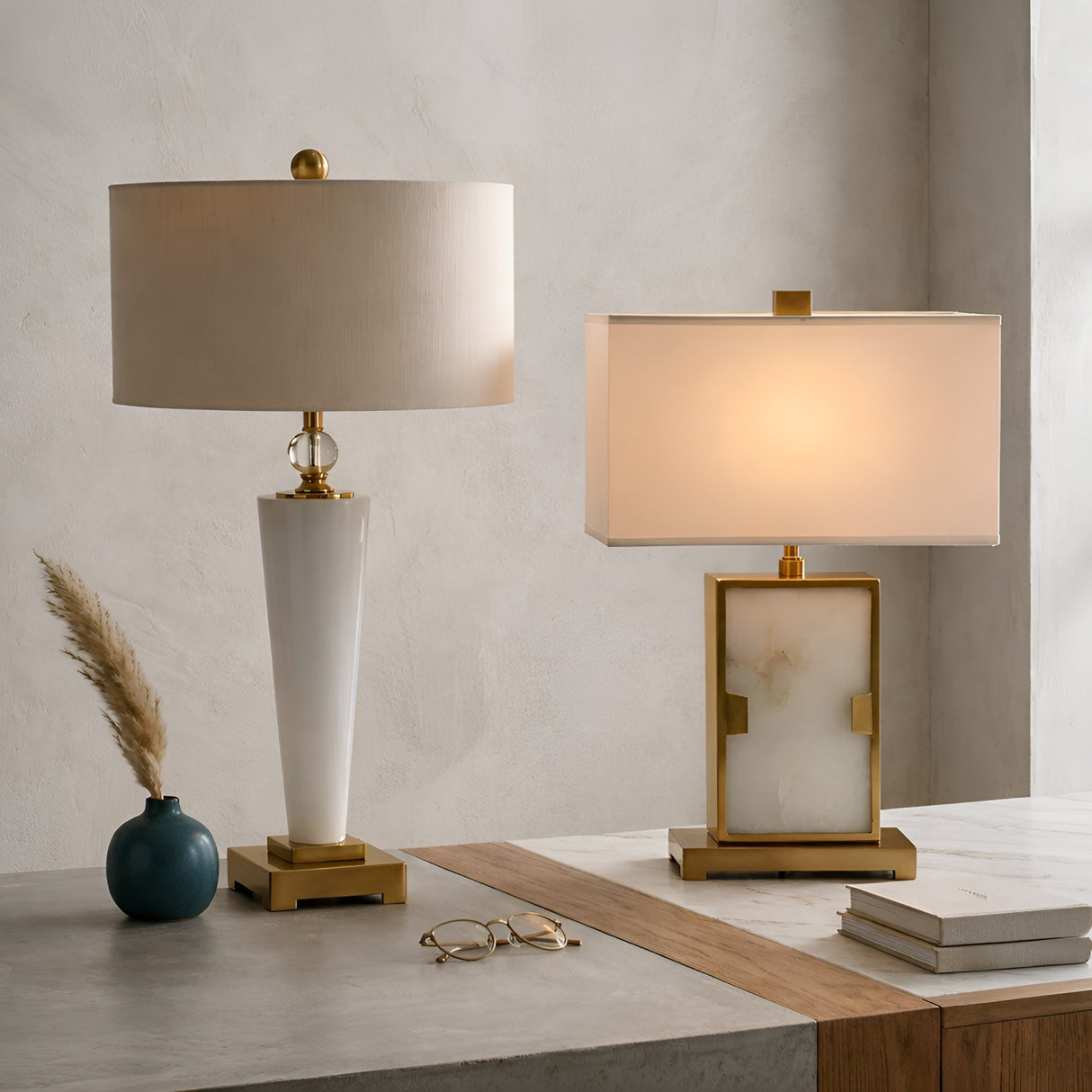 The Aureum 1 Light Refined Silhouette Marble Table Lamp in brass and white sits on a marble surface beside a blue vase with dried grass and eyeglasses, offering a modern touch with its cone base and round shade.