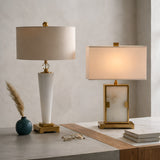 The Aureum 1 Light Refined Silhouette Marble Table Lamp in brass and white sits on a marble surface beside a blue vase with dried grass and eyeglasses, offering a modern touch with its cone base and round shade.