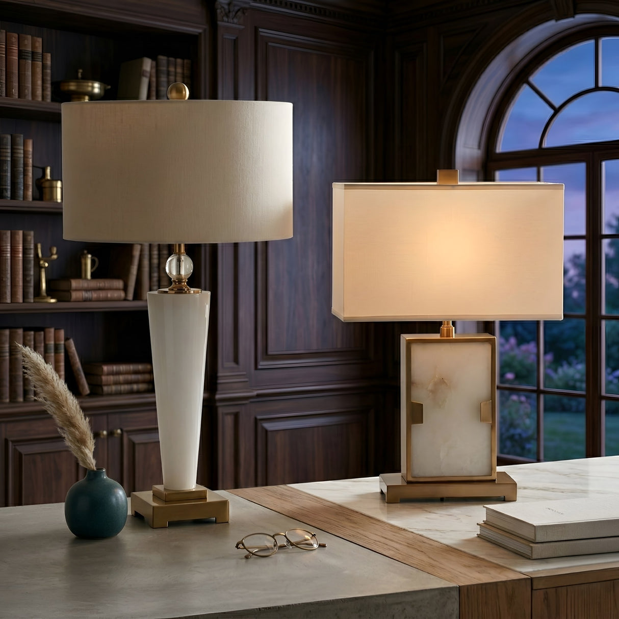 Two Aureum 1 Light Refined Silhouette Marble Table Lamps in brass and white, with white shades, sit on a marble desk in a wood-paneled study. Nearby, bookshelves, an arched window, glasses, and a vase with pampas grass complete the scene.
