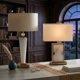 Two Aureum 1 Light Refined Silhouette Marble Table Lamps in brass and white, with white shades, sit on a marble desk in a wood-paneled study. Nearby, bookshelves, an arched window, glasses, and a vase with pampas grass complete the scene.