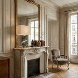 Elegant living room with ornate white fireplace, large gold-framed mirror, Selvarn 1 Light Banded Marble Table Lamp in Brass & Green, decorative vases, and armchair. Tall windows with beige curtains let in natural light on herringbone wood floors.