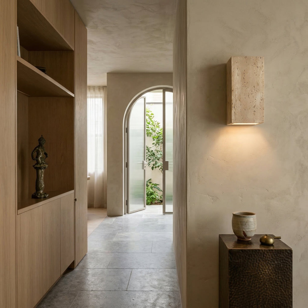 A modern hallway with light wood built-ins, a bronze statue, textured beige walls, the Cavaro LED Rectangular Travertine Wall Light in light beige, a small ceramic-topped table, and an arched glass door to a sunny green courtyard.