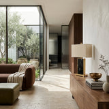 A modern living room has a brown sofa, green ottoman, and wooden sideboard topped with the Valmor 1 Light Block Marble Table Lamp in brass and black. Large glass doors open to a leafy courtyard garden that fills the space with natural light.