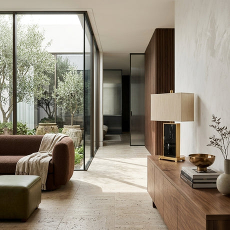 A modern living room has a brown sofa, green ottoman, and wooden sideboard topped with the Valmor 1 Light Block Marble Table Lamp in brass and black. Large glass doors open to a leafy courtyard garden that fills the space with natural light.