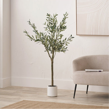 Medium Olive Tree in Pot, 150cm faux tree with green foliage on a bare trunk in a white cylindrical pot, styled in a living room beside a beige armchair.