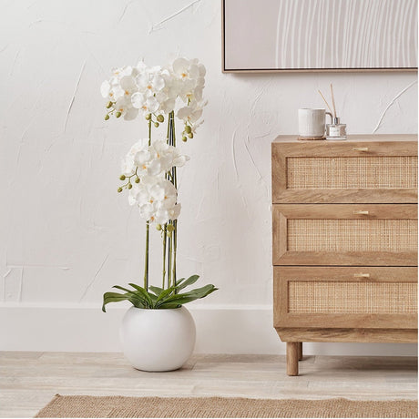 Tall White Orchid in Pot Artificial Plant, 115cm, with layered white blooms and green foliage in a round white ceramic pot, styled beside a wooden cane-front chest of drawers.