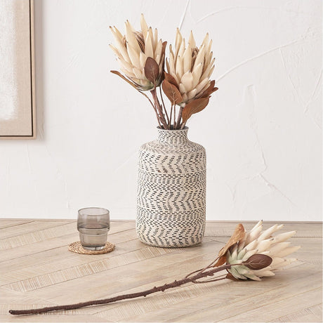 Artificial Protea flowers in natural cream tones displayed in a patterned white and black vase, with a single stem laid on a wooden floor beside a glass tumbler.