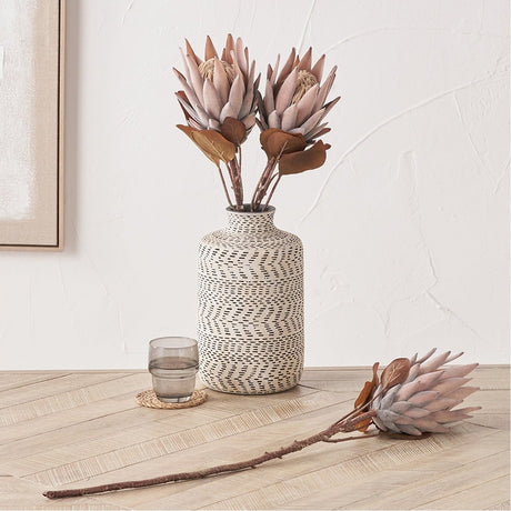 Mulberry Brown Protea artificial flower stems in a dashed cream and black patterned vase on a wooden herringbone floor, with one stem laid beside it, against a plain light wall.