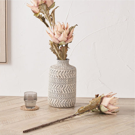 Artificial protea-style stems with pale apricot-pink blooms and green foliage in a patterned cream and black vase, with a matching loose stem laid on a wooden floor beside a grey glass tumbler.