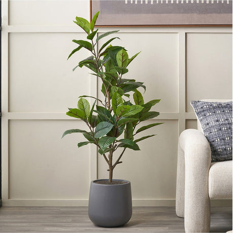 Artificial Magnolia Tree in Pot, 130cm, with green glossy leaves in a matte grey planter, shown in a living room beside a beige sofa and panelled wall.