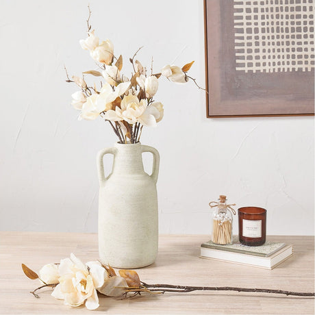 Artificial cream magnolia flower stems arranged in a textured cream ceramic vase with handles, styled on a wooden table beside a loose stem, candle, and books.