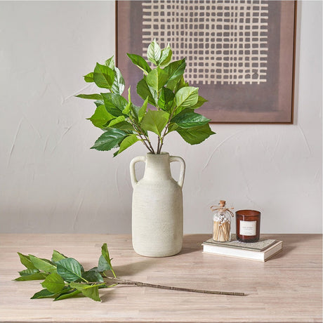Green Hydrangea Leaf artificial foliage stems, 33cm, arranged in a textured cream vase on a wooden sideboard beside a loose stem, candle, and framed wall art.