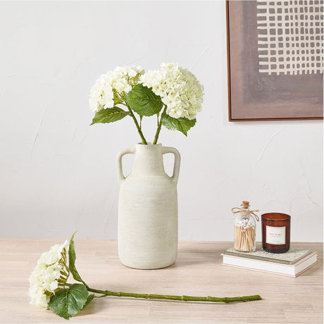White Hydrangea artificial flower stems with green leaves displayed in a textured cream ceramic two-handled vase on a wooden table beside a candle and matchstick jar.