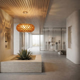A modern, minimalist interior features a succulent in a central planter, smooth beige walls, wooden ceiling panels, glass partitions with artfully lit vases, and the Briller LED Large Suspension Light in gold as a warm focal point.