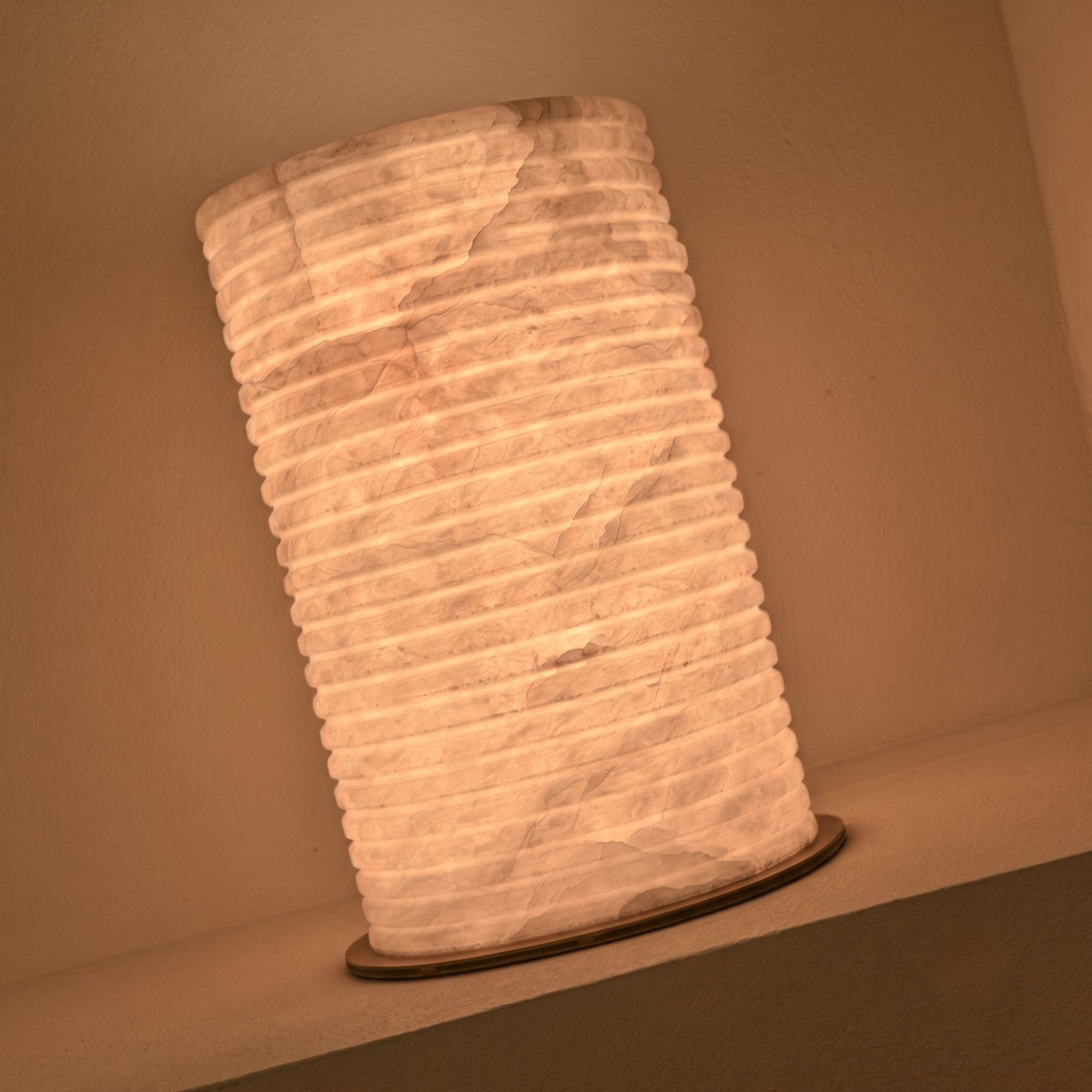 Aurelior Small LED table lamp with ribbed cylindrical alabaster body and brass base disc, glowing warm white in a wall niche setting.