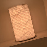 Aurelior Small LED table lamp with ribbed cylindrical alabaster body and brass base disc, glowing warm white in a wall niche setting.