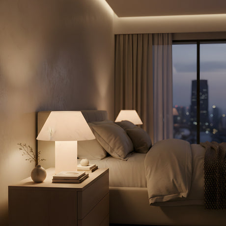 A cozy bedroom at dusk features the Lunessa LED Small Sculpted Mushroom Alabaster Table Lamp – Soft White on the nightstand with stacked books, a small vase, gray pillows on a neatly made bed, and large windows revealing the cityscape.