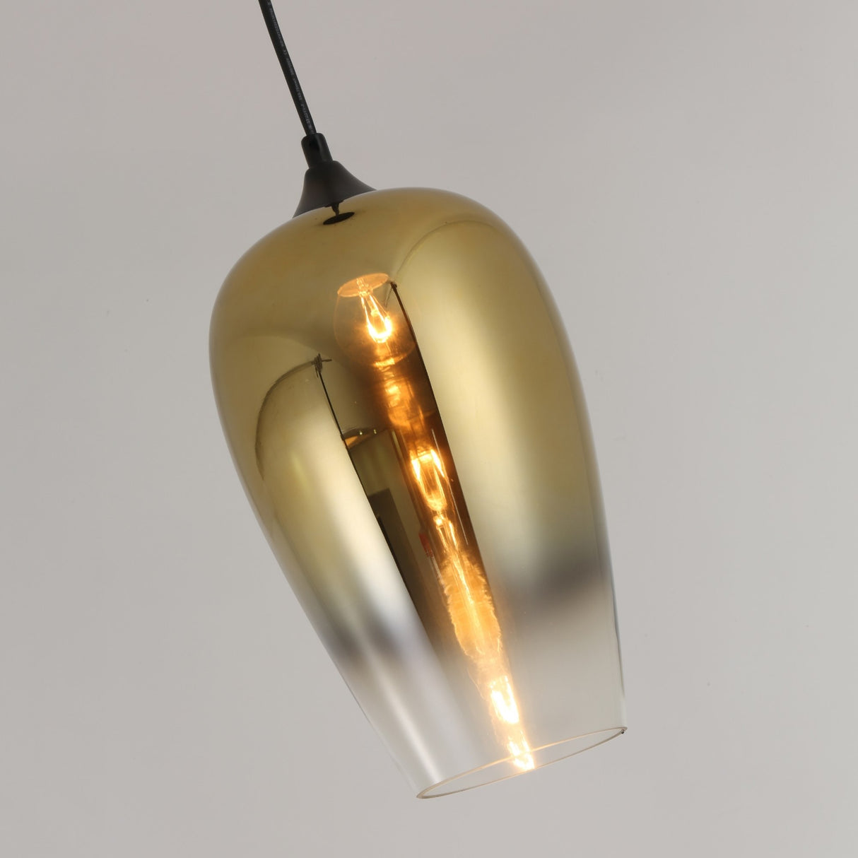 The Slice Glass Pendant Light - Gold features a metallic glass shade suspended by a black cord, casting a warm glow against a light gray background—a striking example of modern handcrafted lighting.