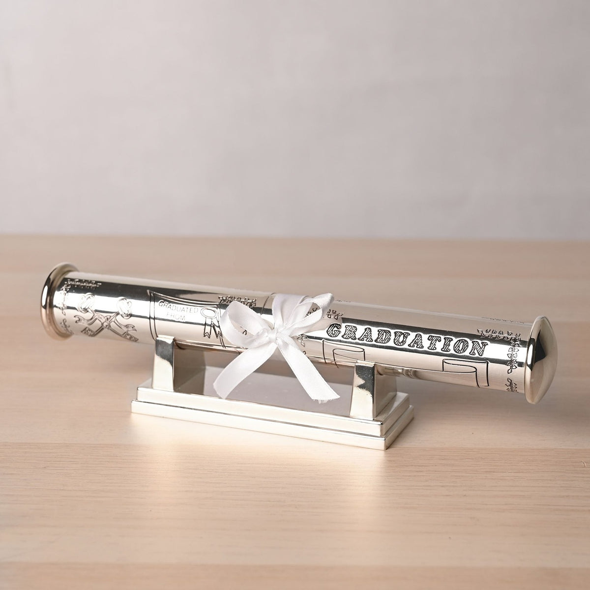 Silverplated graduation certificate holder, a cylindrical tube engraved with celebratory motifs, tied with a white ribbon bow and resting on a matching silver stand on a wooden surface.