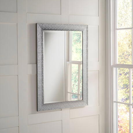 The Eclipso Rectangle Wall Mirror - Silver, featuring its textured silver frame, is gracefully mounted on a white paneled wall. It wonderfully reflects a portion of a window with views of lush trees and a garden, enhancing the allure of the modern interior.