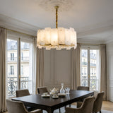 The Caldrin 6 Light Tiered Panel Alabaster Chandelier in Soft White & Brass hangs over a dark dining table with tea service in an elegant room featuring tall windows, cream curtains, ornate moulding, and views of Parisian-style buildings.