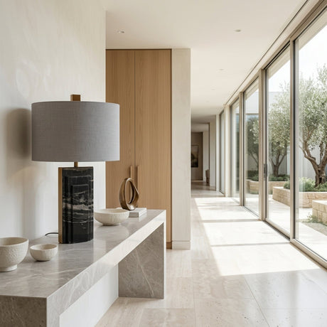 Modern hallway with large windows, a marble-topped console table displaying the Caldrin 1 Light Marble Table Lamp in Brass & Black and ceramic bowls. Sunlight streams in, illuminating the minimalist space with light wood doors and neutral tones.