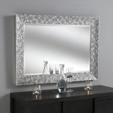 The Astral Rectangle Wall Mirror - Silver, with its ornate frame featuring concentric circles, hangs on a gray wall. Below it, a dark wooden console displays several glass bottles and teardrop-shaped candle holders. The reflections on the mirror's surface shimmer, adding depth to the elegant setting.