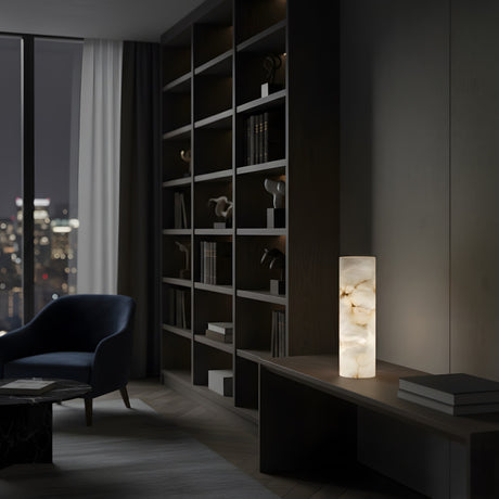 A modern, dimly lit living room with tall bookshelves, a navy armchair, a round table, and the Virelle LED Column Alabaster Table Lamp - Soft White glowing on a wooden console. City buildings shine through the large night window.