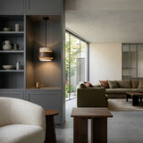 Modern living room with gray built-ins, a white textured armchair, wood tables, a cozy green sofa, and bright garden-facing windows—illuminated by the Virelli LED Drum Travertine Single Pendant Light in Walnut Brown & Light Beige overhead.