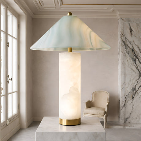 The Veridra 1 Light Conical Shade Alabaster Table Lamp in Soft White & Green, featuring a cylindrical marble base and wide translucent shade, is displayed on a pedestal in an elegant, bright room with marble accents and classic décor.
