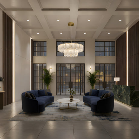 Modern lobby with high ceilings, two navy blue sofas, a round coffee table, the Valeris 28 Light 2 Tier Layard Panel Alabaster Chandelier in Soft White & Brass, green plants, tall windows, wall paneling, and a green marble reception desk on the right. Elegant and well-lit.