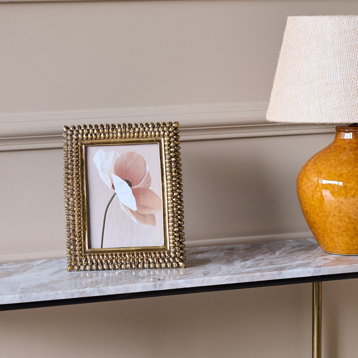 Libra Interiors textured beaded gold resin photo frame displaying a floral print, stood on a marble console table beside an amber glass lamp.
