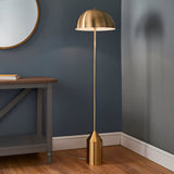 Endon Castford floor lamp in antique brass with a domed shade and cylindrical base, styled beside a wooden console table against a dark blue wall.