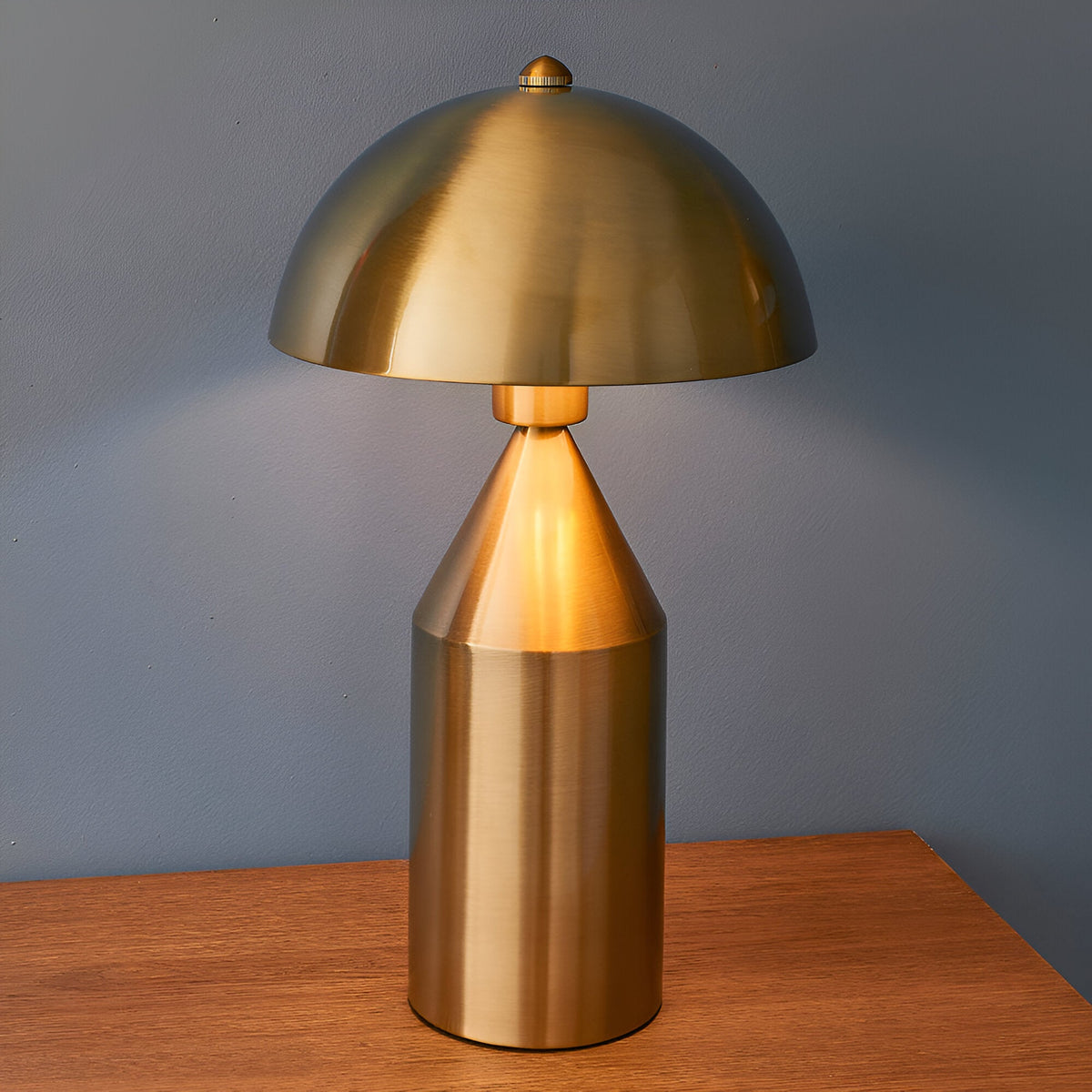Castford Table Lamp in antique brass with a domed metal shade and cylindrical base, shown lit on a wooden surface against a blue wall.