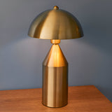 Castford Table Lamp in antique brass with a domed metal shade and cylindrical base, shown lit on a wooden surface against a blue wall.