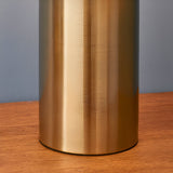 Close-up of the Castford Table Lamp's cylindrical base in antique brass with a brushed metal finish, shown on a wood surface against a grey wall.