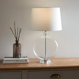 Endon Bramleigh table lamp with clear glass ellipsoid base and white tapered shade on a wooden sideboard beside a vase and books.