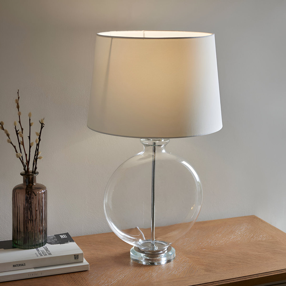 Endon Bramleigh table lamp with clear glass ellipsoid base, nickel stem and white tapered shade, on a wooden console beside books and a vase.
