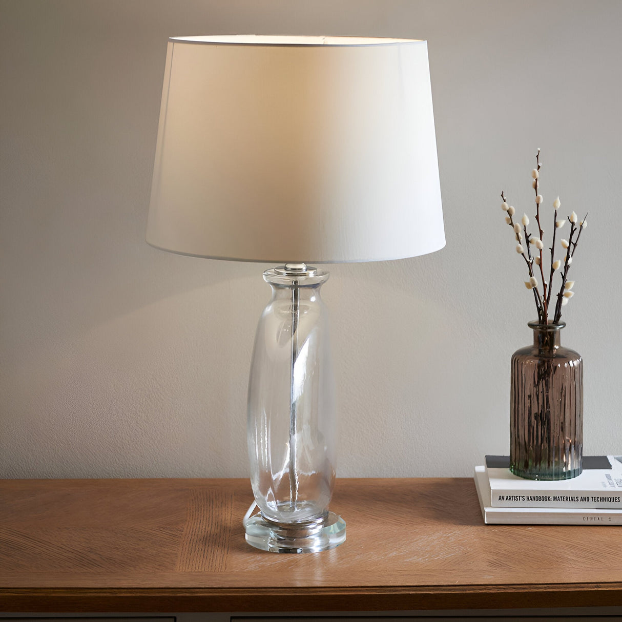 Endon Bramleigh table lamp with a clear glass ellipsoid base, nickel fitting and tapered white shade, lit on a wooden console beside books and a vase.