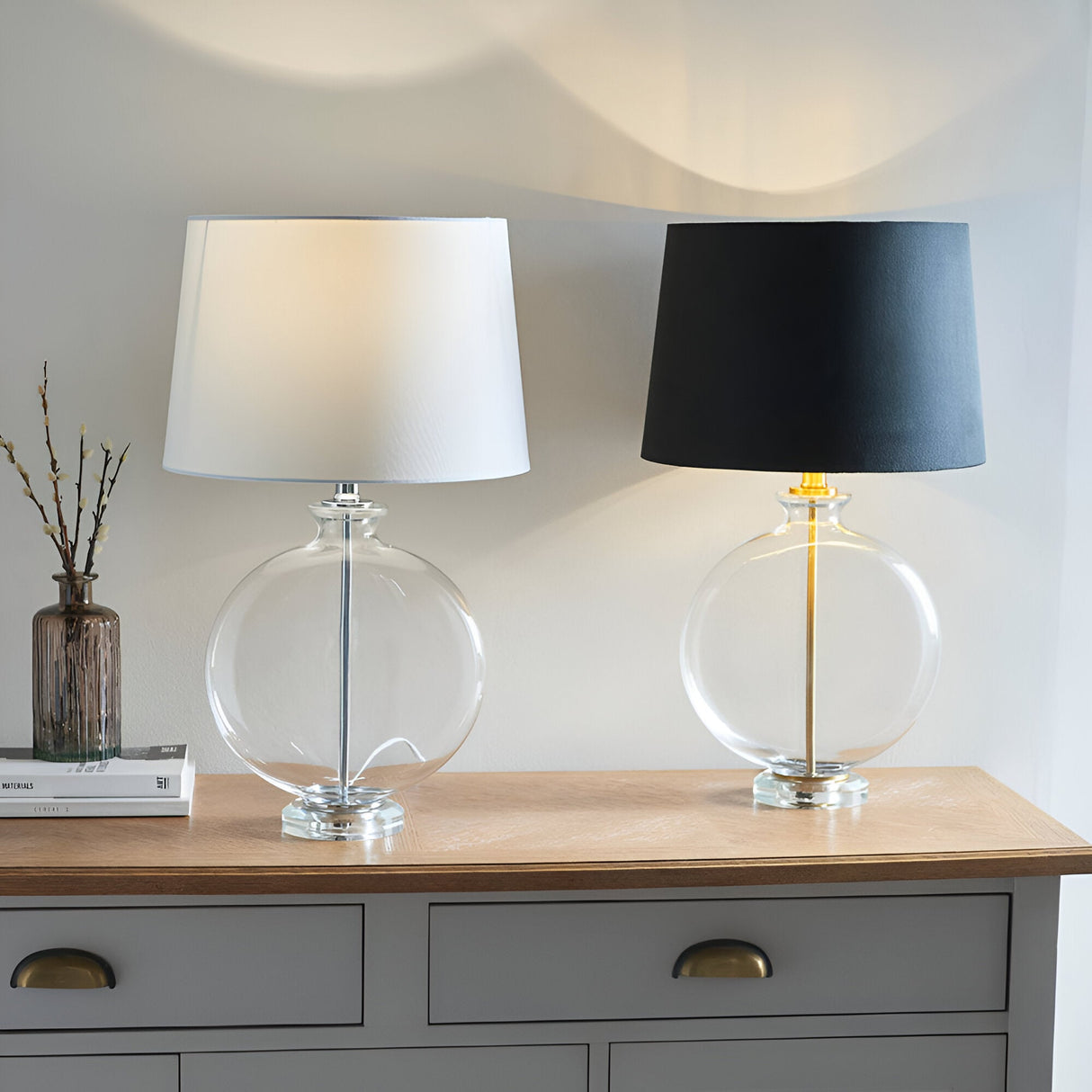 Endon Bramleigh table lamps on a grey sideboard, clear glass ellipsoid bases with nickel and white shade beside brass and black shade.