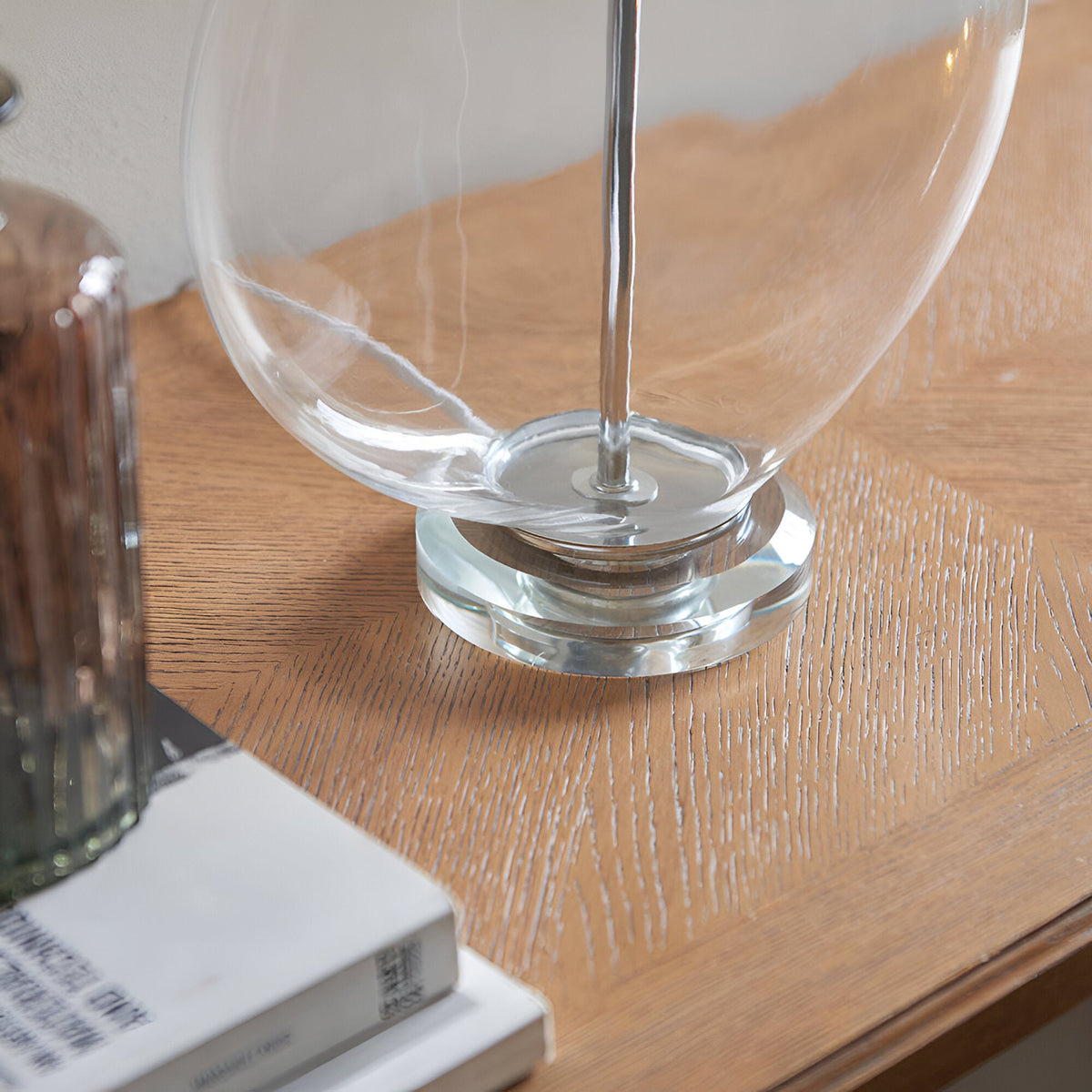 Close-up of the Endon Bramleigh table lamp's clear glass ellipsoid base and polished nickel stem on a wooden console beside books.