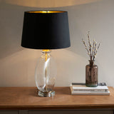 Endon Bramleigh table lamp with clear glass ellipsoid base and black tapered shade with brass interior, on a wooden console beside books and a vase.
