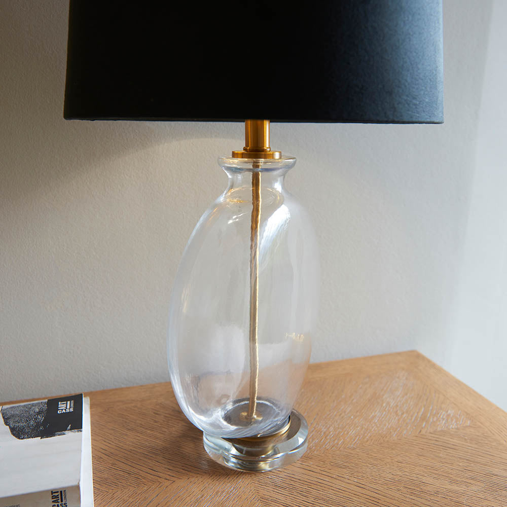 Endon Bramleigh table lamp with clear glass ellipsoid base, antique brass neck and black faux suede tapered shade, on a wooden console beside books.