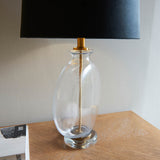 Endon Bramleigh table lamp with clear glass ellipsoid base, antique brass neck and black faux suede tapered shade, on a wooden console beside books.