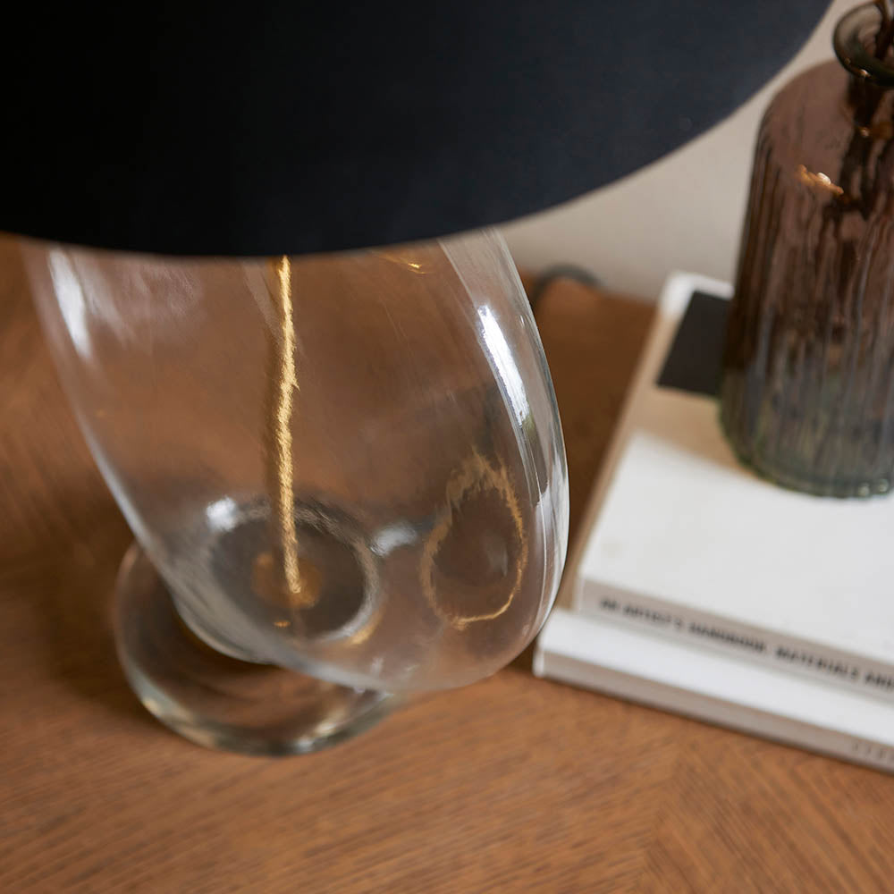 Close-up of the Endon Bramleigh table lamp's clear glass ellipsoid base and black shade on a wood surface beside stacked books and a brown glass vessel.